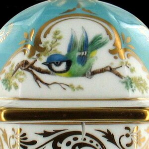 May include: A porcelain box with a blue and gold lid. The lid features a hand-painted bluebird perched on a branch with green leaves. The box has a hexagonal shape and intricate gold detailing.