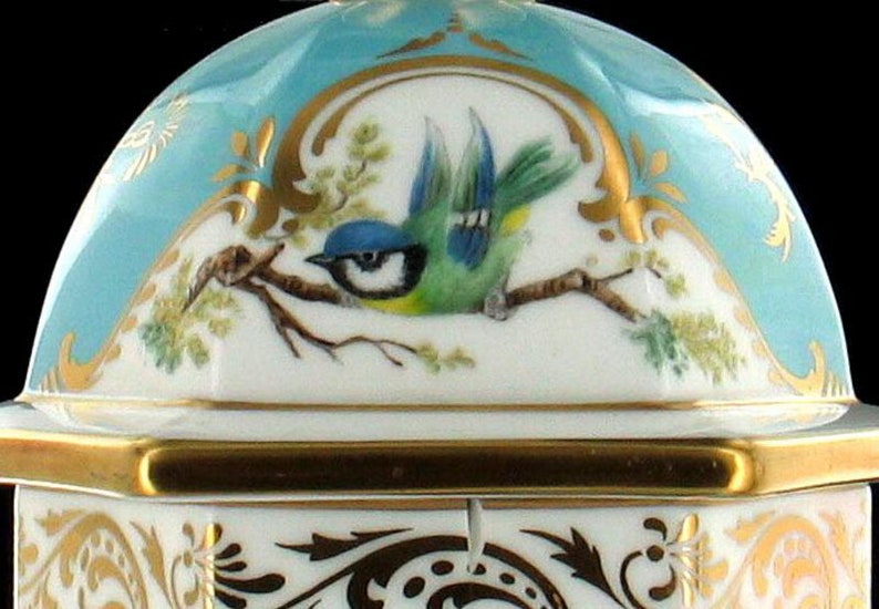 May include: A porcelain box with a blue and gold lid. The lid features a hand-painted bluebird perched on a branch with green leaves. The box has a hexagonal shape and intricate gold detailing.