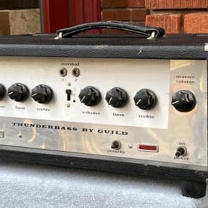 May include: A black Guild Thunderbass guitar amplifier with a silver faceplate and multiple knobs. The amplifier has a built-in handle and a red light on the front.