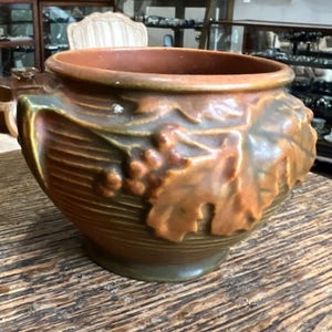 May include: A brown and green glazed ceramic planter with a raised design of leaves and grapes. The planter has two handles on the sides.