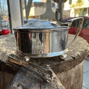 Puede incluir: Un azucarero de plata con tapa y un par de pinzas para azúcar a juego. El bol tiene una forma redondeada y un asa decorativa. Las pinzas son de plata y tienen un diseño sencillo.