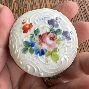May include: A white porcelain trinket box featuring a floral design. The box is decorated with hand-painted flowers in shades of pink, red, blue, and purple. The box has a white background with a delicate, embossed pattern.