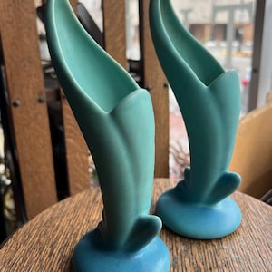 Puede incluir: Dos jarrones de cerámica turquesa con un diseño en forma de tulipán. Los jarrones tienen una parte superior acampanada y una base redondeada. Los jarrones están sobre una mesa de madera. Los jarrones son de color azul verdoso claro.