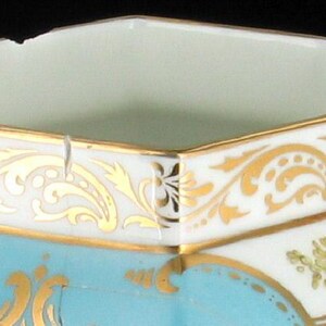 May include: A hexagonal porcelain sugar bowl with a light blue glaze and gold trim. The bowl has a floral design and a gold rim.