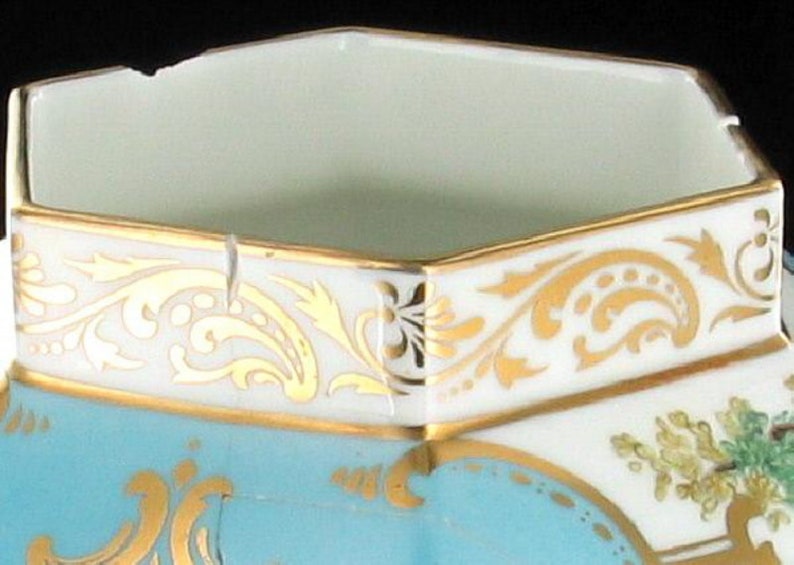 May include: A hexagonal porcelain sugar bowl with a light blue glaze and gold trim. The bowl has a floral design and a gold rim.