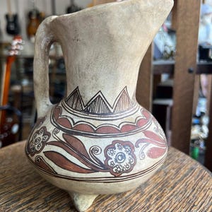 May include: A beige ceramic pitcher with a handle and a spout. The pitcher is decorated with brown geometric and floral patterns. It has three small feet and is sitting on a wooden surface.