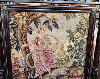 Può includere: Un parascintille in legno scuro con un pannello di arazzo che raffigura una donna in un abito rosa in piedi in un bosco con un agnello bianco. L'arazzo è circondato da una cornice in legno scuro con intagli decorativi.