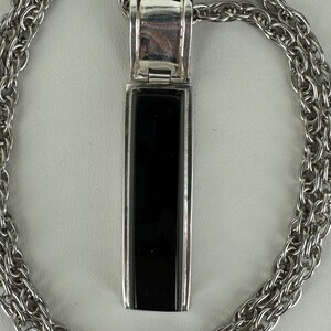 May include: A silver-toned necklace with a rectangular black pendant. The pendant is framed in silver and hangs from a matching chain. The chain has a woven design. The necklace is displayed on a white surface.