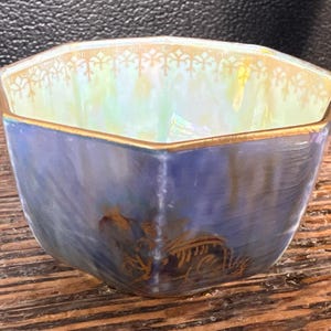 May include: An octagonal bowl with a blue exterior and gold trim. The interior has an iridescent finish. The bowl's design includes gold accents and a decorative pattern along the rim. The bowl is sitting on a wooden surface.