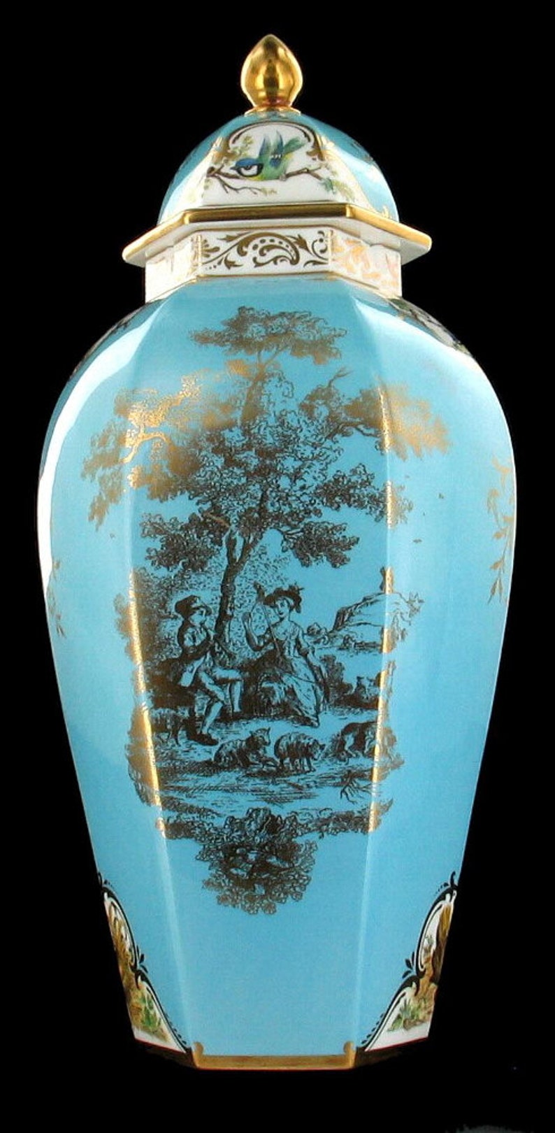 May include: A blue and gold porcelain vase with a lid. The vase is decorated with a pastoral scene of two figures and sheep in a landscape. The lid is decorated with a bird.
