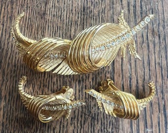 以下が含まれることがあります： 金色のジュエリーセット。ブローチとイヤリングのセットで、羽をモチーフにしたデザインで、クリアなラインストーンが装飾されています。木製の表面に展示されています。