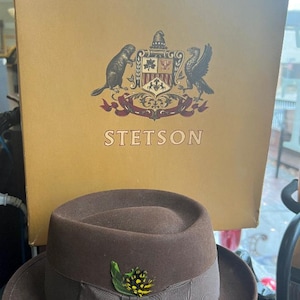 Op de afbeelding: Een bruine wollen fedora hoed met een bruin lint en een klein groen en geel bloemetje. De hoed ligt op een bruin oppervlak voor een doos met het Stetson-logo.