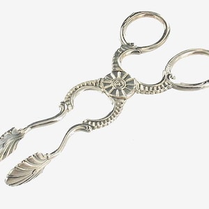 May include: Silver sugar tongs with an ornate design. The tongs have a decorative floral pattern and a rounded handle.