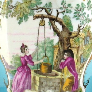 May include: A vintage porcelain vase with a blue and gold design. The vase features a painted scene of a woman and a man standing by a well with a tree and a small house in the background. Two sheep are grazing in the foreground.