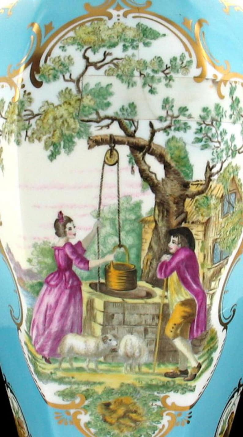 May include: A vintage porcelain vase with a blue and gold design. The vase features a painted scene of a woman and a man standing by a well with a tree and a small house in the background. Two sheep are grazing in the foreground.