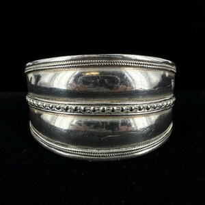 Puede incluir: Un brazalete de plata con una banda ancha y redondeada y tres bordes decorativos. Los bordes están hechos de pequeñas cuentas y un diseño de cuerda.