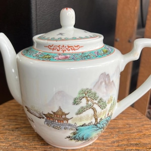 May include: White porcelain teapot with a lid, featuring a hand-painted landscape scene with a pagoda, mountains, and a tree. The lid has a red finial and decorative trim. The handle and spout are curved.