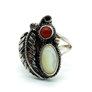 May include: A silver ring with a white oval stone and two red round stones. The ring has a detailed design with a leaf-like pattern.