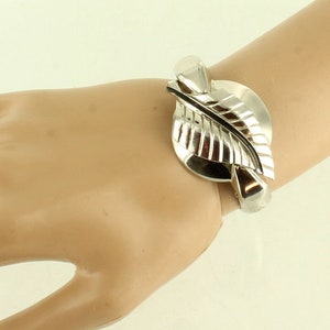 May include: A silver bracelet featuring a leaf design. The bracelet is open and has a hinged clasp.