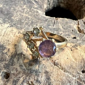 May include: A gold ring with a large purple gemstone and smaller clear rhinestones set in a triangular shape.