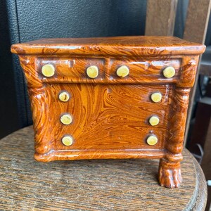 May include: A miniature ceramic dresser with a marbled brown and orange finish. The dresser has two drawers and six yellow knobs. The legs are shaped like claw feet. A decorative accent piece.