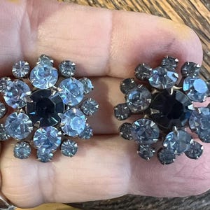 Puede incluir: Un par de pendientes de botón de estilo vintage. Cada pendiente presenta una piedra central azul oscuro rodeada de pedrería azul claro y gris, dispuesta en un patrón floral. Los pendientes están engarzados sobre una base de metal plateado.