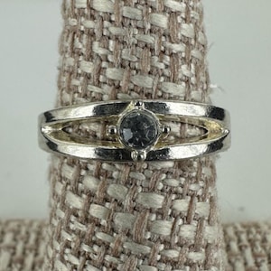 Op de afbeelding: Zilverkleurige ring met een centrale, ronde, grijze edelsteen. De ring heeft een gespleten bandontwerp met kleine, decoratieve accenten. De ring wordt gepresenteerd op een getextureerd, neutraal gekleurd oppervlak.