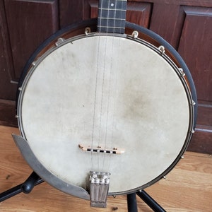 May include: A vintage banjo with a round, white head and a wooden neck. The banjo has a metal tuning peg and a metal bridge.