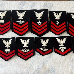 以下が含まれることがあります： ヴィンテージの海軍パッチのコレクション。各パッチは黒色で、上部に白いワシのエンブレムと赤いシェブロンが付いています。交差したツールやアンカーなど、さまざまなサービスシンボルがワシの下に表示されています。