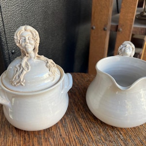 May include: A white ceramic sugar bowl with a lid and a matching creamer. Both pieces feature a sculpted face on the top. The sugar bowl has handles on either side. The creamer has a spout.