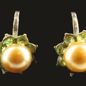 May include: A pair of earrings featuring a large, round, golden-colored pearl at the center, surrounded by a cluster of small, light green gemstones. The earrings have a silver-colored hook for pierced ears. The background is black.