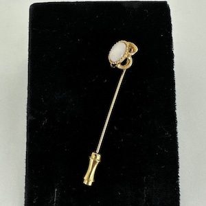 Op de afbeelding: Een gouden stickpin met een witte ovale steen gezet in een gouden filigraanontwerp.