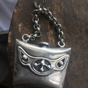 May include: A silver charm in the shape of a small, rectangular purse with a chain attached. The purse has a decorative design with small circles and a central design.