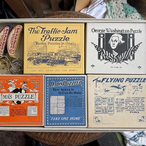 May include: A collection of vintage puzzles and games in a white box. The box contains six puzzle boxes with various graphics and text, including "The Traffic Jam Puzzle" and "George Washington Puzzle". Also included are two "Roly Poly Puzzle" toys.