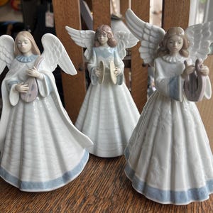 May include: Three porcelain angel figurines. Each angel wears a white gown with a light blue trim and holds a musical instrument. The angels have white wings and are standing on a wooden surface.