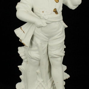 Może przedstawiać: Biała porcelanowa figurka przedstawiająca młodego chłopca w kapeluszu trzymającego kij. Figurka stoi na podstawie z ozdobnym wzorem.