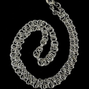May include: A silver-colored necklace with a double-strand chainmail design. The necklace is made of small, interlocking rings, creating a textured and intricate pattern. The clasp is visible at the top, and the necklace is displayed against a black background.