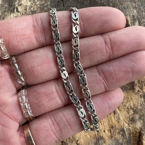 May include: Two silver-colored chain necklaces with a repeating swirl design are displayed. The chains are held in a hand, with a gold ring and a clear ring visible. The background is a weathered wooden surface.