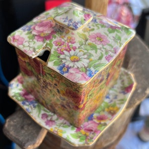 Puede incluir: Un azucarero cuadrado de cerámica con tapa, con un patrón floral en tonos de rosa, amarillo, azul y verde. El tazón y la tapa están adornados con detalles dorados. El tazón se asienta sobre un platillo a juego.