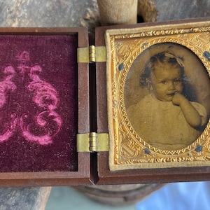 May include: A vintage wooden photograph case with a hinged lid. The case is lined with burgundy velvet and features a decorative embossed design. The case holds a black and white photograph of a young child with a bow in their hair, looking at the camera with a finger in their mouth.