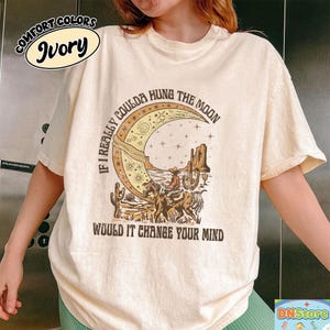 May include: Ivory-colored t-shirt with a graphic featuring a cowboy riding a horse under a crescent moon. The text reads "If I Really Coulda Hung The Moon, Would It Change Your Mind?" The shirt also has the words "Comfort Colors" and "Ivory".