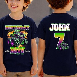 K&ouml;nnte beinhalten: Marineblaues Geburtstags-T-Shirt mit einem Monster-Truck-Motiv in Gr&uuml;n und Lila. Auf der Vorderseite des Shirts steht "BIRTHDAY BOY". Auf der R&uuml;ckseite des Shirts stehen der Name "JOHN" und die Zahl "7".
