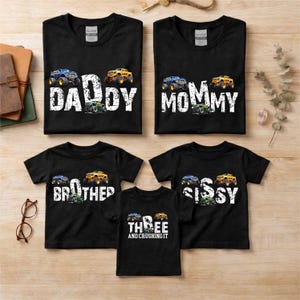 以下が含まれることがあります： モンスター トラックのグラフィックとテキストがプリントされた黒いTシャツ。「Daddy」、「Mommy」、「Brother」、「Sissy」、「Three and Crushing It」と書かれています。 モンスター トラックは青、黄色、緑色です。