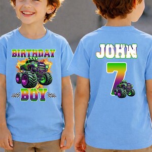 K&ouml;nnte beinhalten: Zwei hellblaue T-Shirts mit einem Monster-Truck-Motiv. Auf einem Shirt steht "BIRTHDAY BOY" und auf dem anderen "JOHN 7". Die Shirts werden von einem jungen Mann getragen, der khakifarbene Shorts tr&auml;gt.