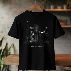 May include: Black t-shirt featuring a distressed white graphic of a guitar. The text "PLAYED UNTIL IT BROKE" is printed below the guitar image. The shirt is displayed on a wooden hanger.