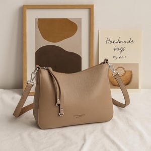 May include: A taupe-colored shoulder bag with a long strap and silver hardware. The bag has a rounded shape and a textured surface. The text "CONGA & LIRIOUM" is printed on the front. The background includes a framed abstract art print and a sign that reads "Handmade bags by azir."