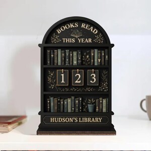 May include: A black wooden bookshelf-shaped calendar with the text "BOOKS READ THIS YEAR" at the top. The calendar displays the numbers "1 2 3" and the text "HUDSON'S LIBRARY". The shelves are filled with miniature books.