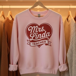 May include: Pale pink sweatshirt featuring a maroon heart design with the words "Mrs. Linda TEACHER" in white script. The sweatshirt has long sleeves and a crew neck. The garment is hanging on a wooden hanger.