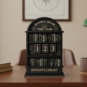 May include: Black wooden bookshelf-shaped calendar with the text "BOOKS READ THIS YEAR" and "HUDSON'S LIBRARY". The calendar displays the numbers 123. The bookshelf is filled with miniature book replicas.