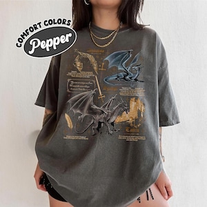 May include: A grey t-shirt with a dragon graphic. The shirt features illustrations of dragons with text and the words "Comfort Colors" and "Pepper" in a circular design.
